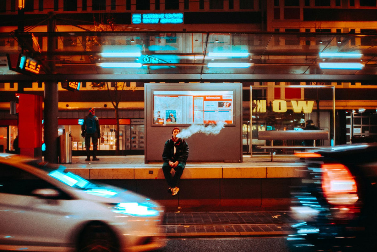 A man sits at a tram stop at night blowing out a cloud of vape while traffic blurs past