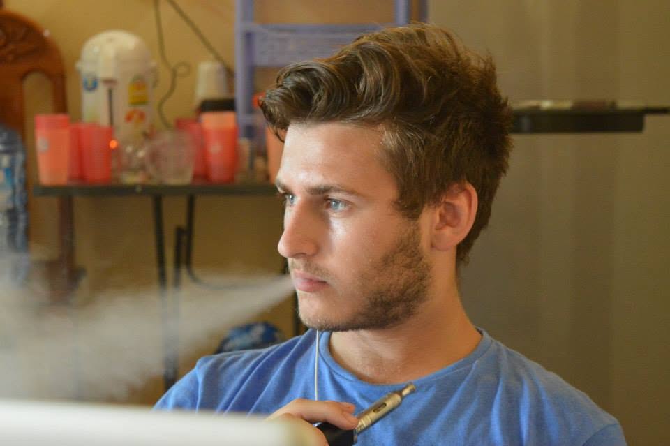 A young man with styled brown hair and a light beard, wearing a blue t-shirt, holding an electronic vape device near his mouth while exhaling a visible plume of white smoke in an indoor setting.